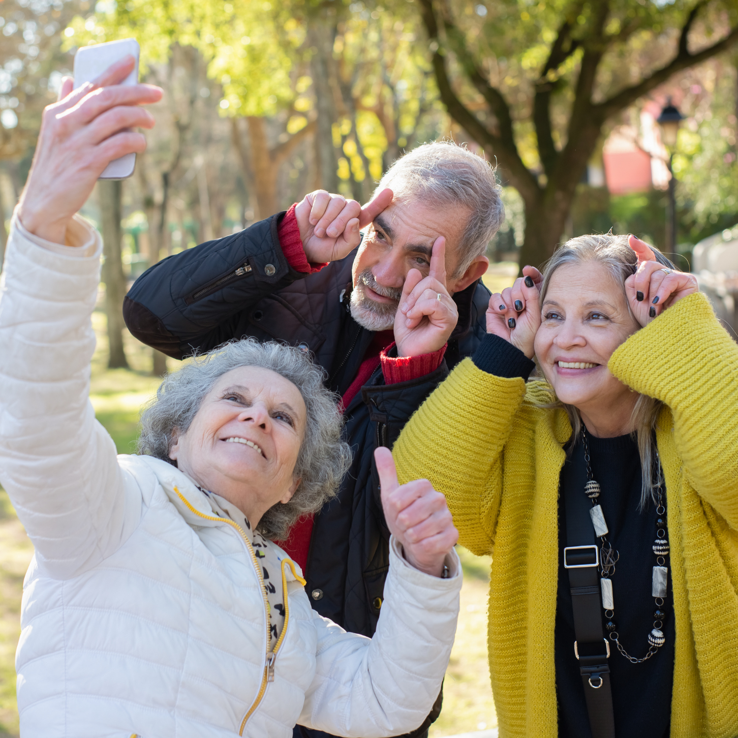 Three smiling older adults take a selfie together outdoors using a smartphone.