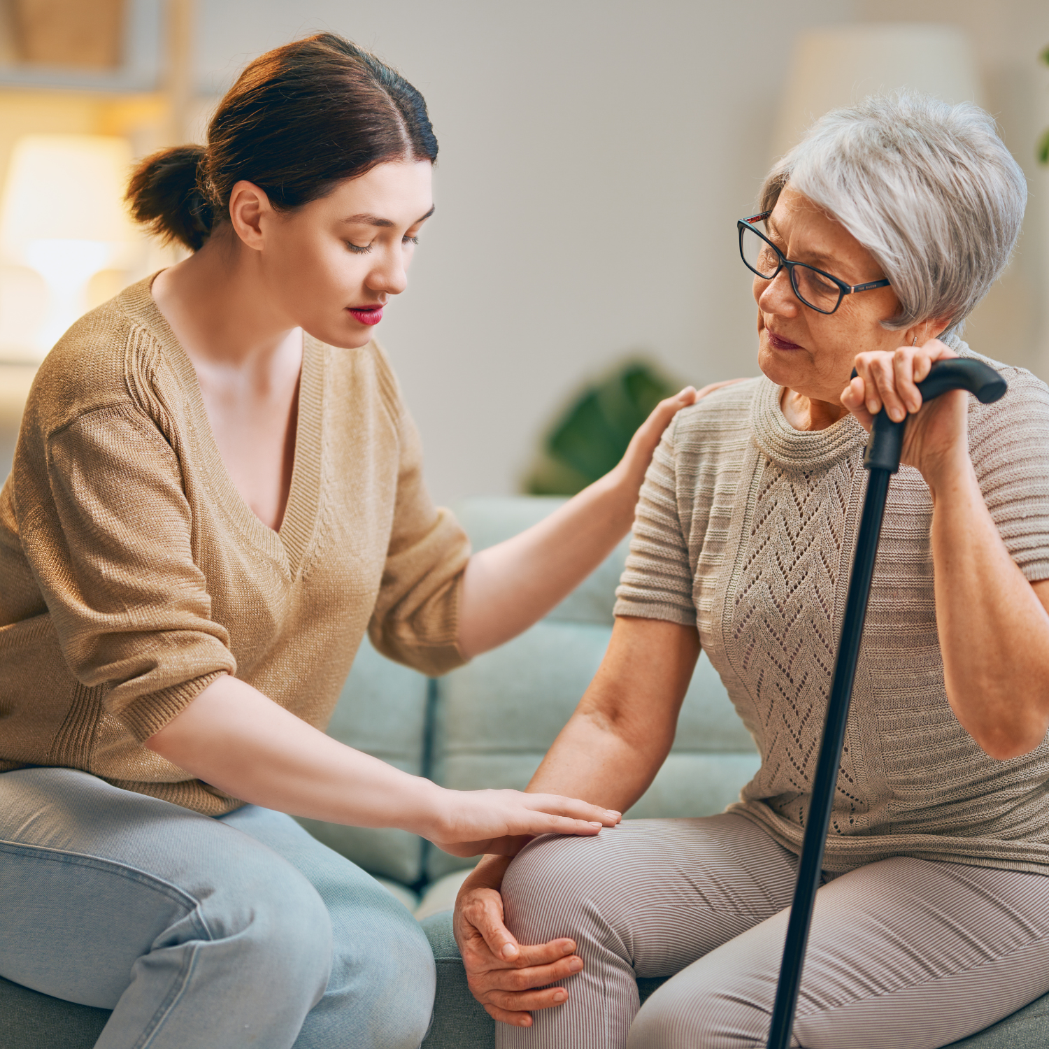 A younger woman comforts an older woman who holds a cane as they talk together on a sofa at home.